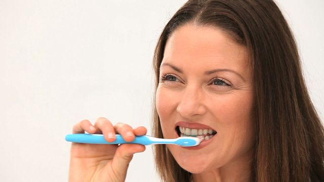 Woman is engaged in oral hygiene using a blue toothbrush pressing softly against teeth while smiling slightly against a simple white backdrop. Perfect for illustrating dental care concepts, health blog posts, teaching materials on personal hygiene, or promoting dental products highlighting routine care.