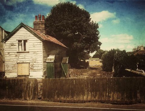 Weathered wooden cottage standing by roadside with overgrown yard and vintage summer light