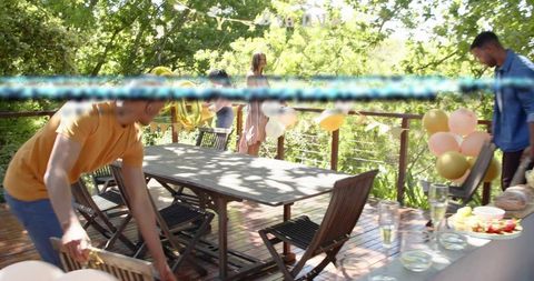 Friends arranging outdoor birthday party on wooden deck with pastel balloons and dining table