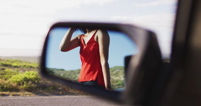 A woman captured in the side mirror of a car, wearing a red top. The image is set on a road with a scenic, green landscape. Use this image for travel, fashion, and lifestyle designs, highlighting themes of journey, contemplation, and scenic beauty.