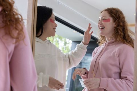 Friends applying pink under-eye patches laughing at vanity in hoodies