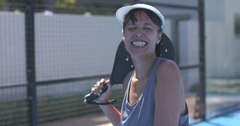 Joyful woman laughing while holding paddle on outdoor blue court during sunny match with visor
