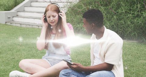 Teen friends sharing headphones on lawn with smartphone and magical light flare
