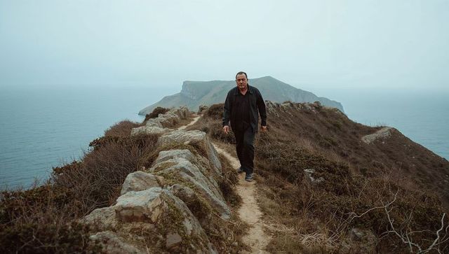 Solitary man walking narrow cliff path on windswept coastal headland