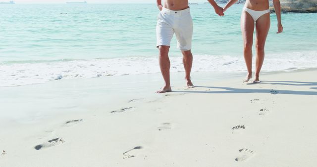 A Caucasian couple enjoys a romantic walk along a sandy beach, with copy space. Their footprints trail behind them, symbolizing a shared journey and intimate moments together.