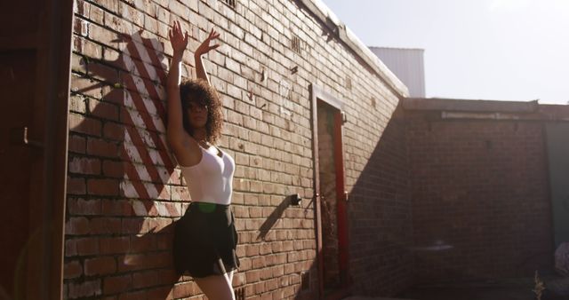 Woman striking dance pose against brick wall on a sunny day, wearing white top, black skirt, and sunglasses. Ideal for use in hip-hop culture promotion, dance school advertisements, urban lifestyle visuals.
