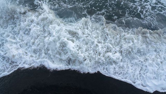 Turbulent surf breaking over black sand shoreline from overhead drone, foamy waves churning