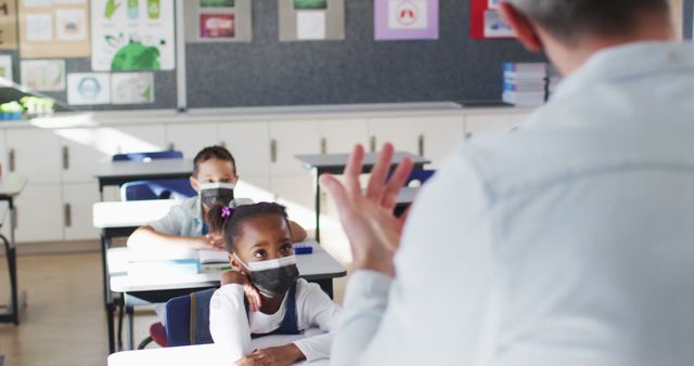 Diverse teacher conducting lesson in classroom with children, all wearing face masks. children in primary school during coronavirus covid 19 pandemic.