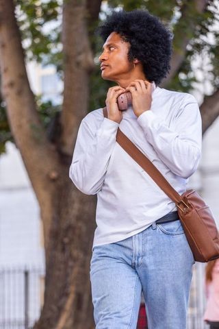 African man standing on urban sidewalk holding pink headphones and brown crossbody bag