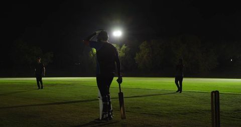 Night cricket batter standing under floodlight holding bat wearing pads helmet on grass field