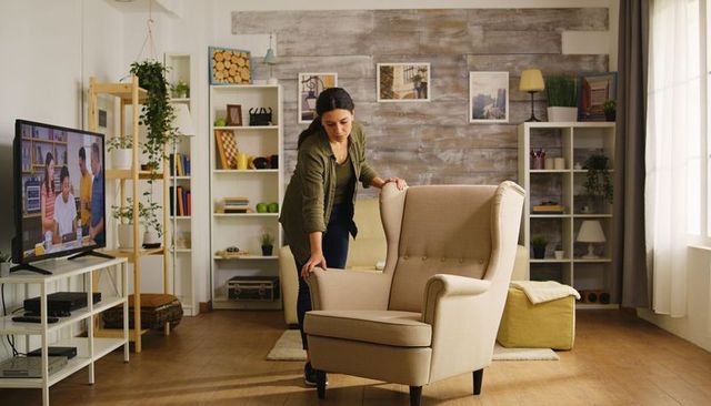 Woman rearranging beige wingback armchair in bright living room with plants and ottoman