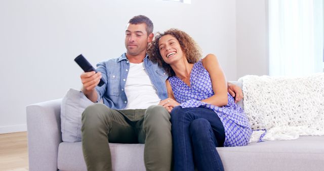 This image shows a happy couple sitting on a couch in their living room, watching TV together. They are enjoying their leisure time, exuding warmth and comfort. Perfect for use in advertisements related to home entertainment, family bonding, or lifestyle blogs.