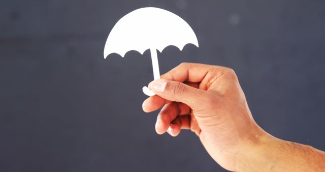 A Caucasian hand is holding a small, white paper cutout of an umbrella, with copy space. It represents creativity or the concept of protection and insurance in a minimalist style.