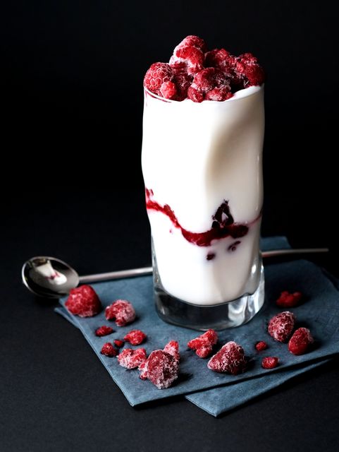 Showcasing a tall glass filled with creamy yogurt topped with frozen raspberries, this image represents a healthy and refreshing dairy dessert. Ideal for nutrition blogs, recipe websites, or advertisements promoting yogurt or dairy products. The contrast against the dark background draws attention to the vibrant colors and textures, making it perfect for culinary magazines and health-focused content.