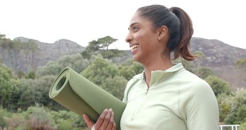 Asian woman smiling and holding yoga mat on mountain trail for outdoor wellness and fitness