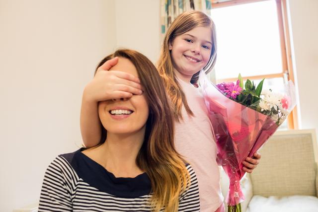 This image shows a young girl surprising her mother with a bouquet of flowers in a cozy living room. The mother is smiling with her eyes covered by her daughter's hand, creating a heartwarming and joyful moment. This image can be used for Mother's Day promotions, family-oriented advertisements, parenting blogs, or any content celebrating family love and special moments.