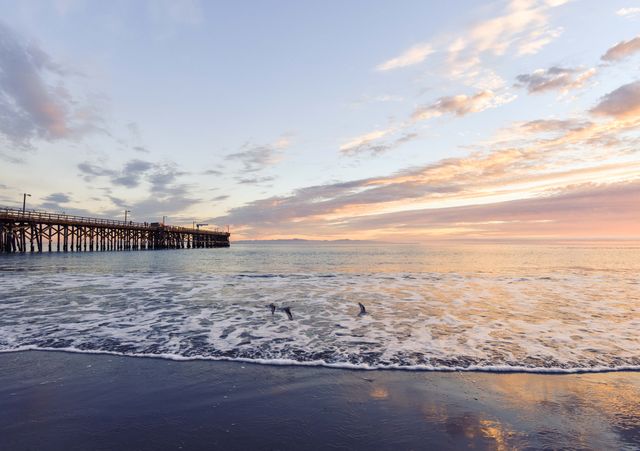 Pier extending into sea during tranquil sunset. Gentle waves breaking onto beach, creating relaxing atmosphere. Ideal for illustrating travel destinations, relaxation, and nature scenes. Perfect use for travel magazines, desktop wallpapers, and calming themed projects.