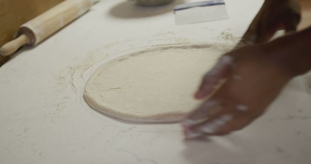 Hands preparing pizza dough on a floured surface in kitchen. Surrounding items like rolling pin indicate active baking process. Perfect for content about homemade cooking, pizza recipes, culinary tutorials, and bakery advertisements.