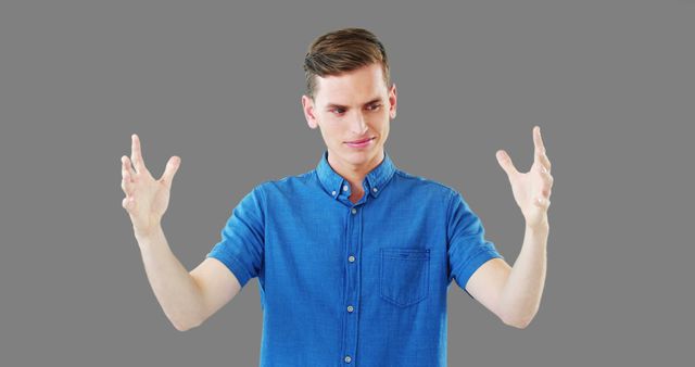Young man in a blue shirt gesturing with both hands against a gray background. Ideal for use in communication themes, corporate training materials, or psychological studies on body language and expressions. Can be used to illustrate concepts such as discussion, explanation, or describing size and scale.