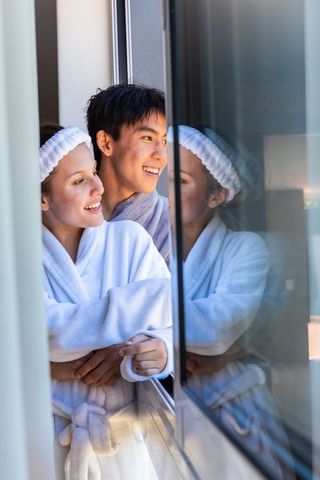 Couple embracing on balcony in plush white robes enjoying spa morning and glass reflection