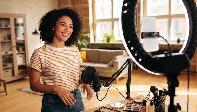 Smiling creator recording makeup tutorial with ring light and microphone in home studio
