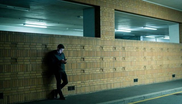 Man leaning on brick wall at night scrolling smartphone in moody urban underground parking