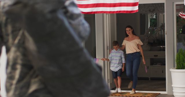 Caucasian male soldier greeting son and wife in garden with american flag hanging outside house. soldier returning home to family.