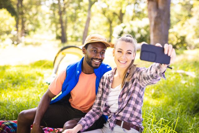 Diverse couple taking selfie with tent in park on sunny day. Spending quality time, lifestyle and camping concept.
