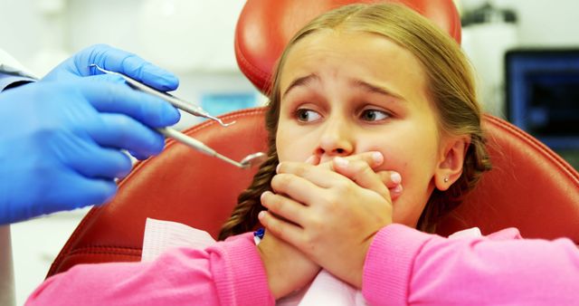 Young girl appears frightened while covering her mouth during a dental checkup. Pediatric dentist and nurse wearing blue gloves, holding dental tools, attempting to ease her anxiety. Suitable for topics related to pediatric dentistry, dental anxiety in children, oral health, medical consultations, dental care.