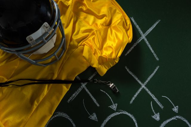 Close-up of American football gear including a yellow jersey, helmet, and referee whistle placed on a green board with a strategy diagram drawn in chalk. Ideal for use in sports-related content, coaching materials, team preparation guides, and articles on American football tactics.