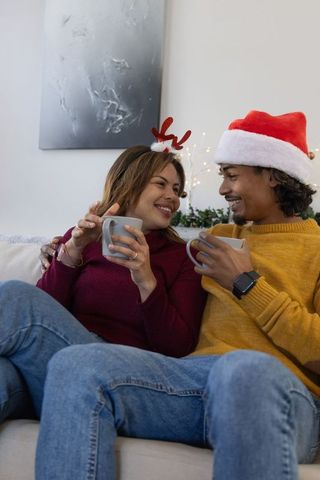 Diverse couple sipping hot drinks on couch wearing santa hat and reindeer headband cozy holiday