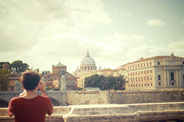 A tourist is photographing the iconic St. Peter's Basilica in Rome. The photo captures the historical buildings and cloudy sky, giving a timeless look. This image is perfect for travel blogs, Rome tourism promotions, and cultural articles.