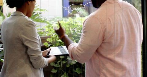 Creative team brainstorming budget strategy on glassboard with tablet and indoor greenery