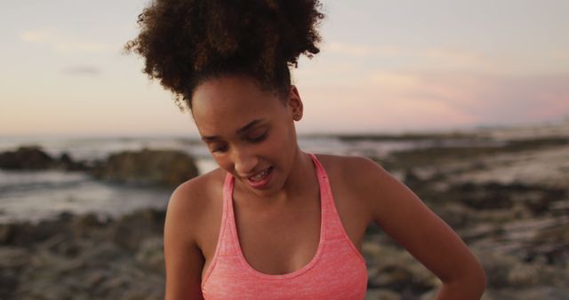 Young Black woman wearing pink sports top, enjoying time at rocky beach during sunset. Great for fitness, nature, and lifestyle themes, as well as beach and outdoor activities.