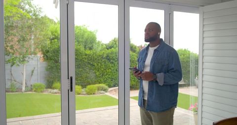 African man standing by sliding glass doors using smartphone and earbuds with garden patio view