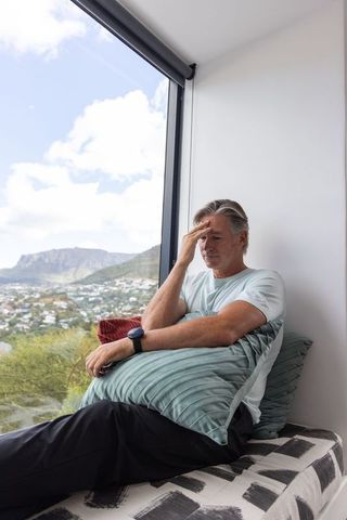 Mature man resting on window seat holding pillow, gazing contemplative over hillside view