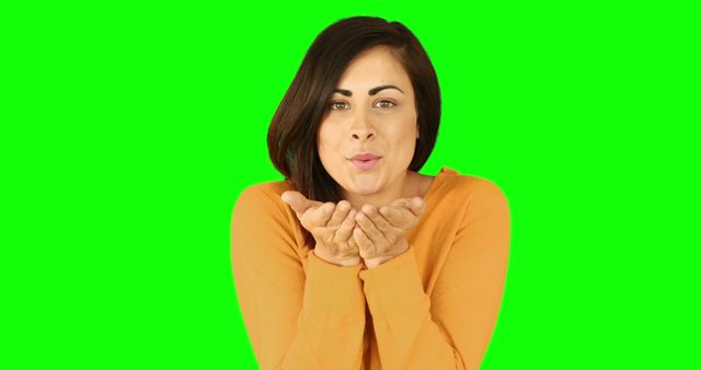 Woman wearing orange sweater blowing kiss gesture on green screen background. Perfect for advertisements, greeting card designs, expressing affection and love in visual content, or use in promotional materials. Green screen background allows for easy background modification in post-production.