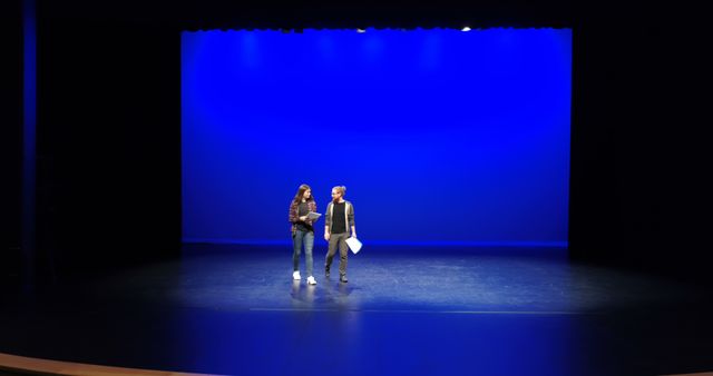 Two people are standing on a stage with a vibrant blue backdrop, engaging in a discussion with copy space. They appear to be actors or directors rehearsing or discussing a scene in a theater setting.