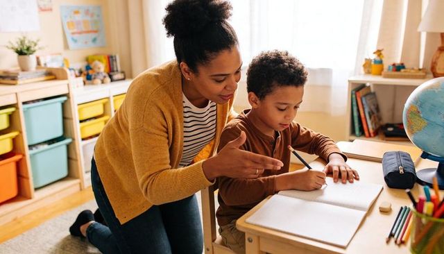 Mother kneeling guiding son writing at desk homeschool tutoring warm family learning moment