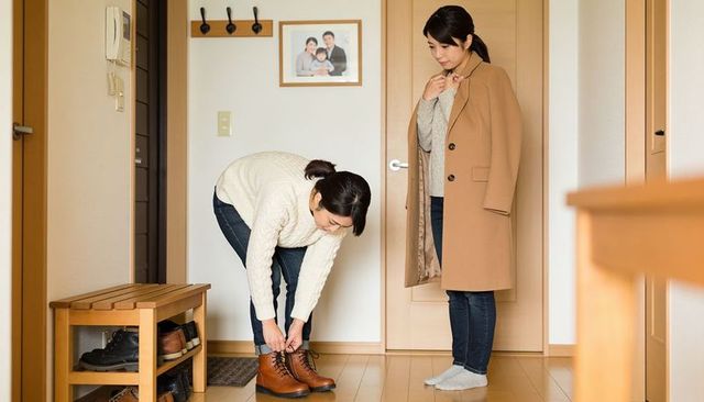 Young woman tying leather boots in home entryway while friend wearing camel coat watches