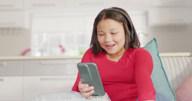 Girl sitting on sofa in stylish living room, engaging in online conversation over smartphone. Perfect for themes of modern technology, children's activities, digital communication, and family home life.