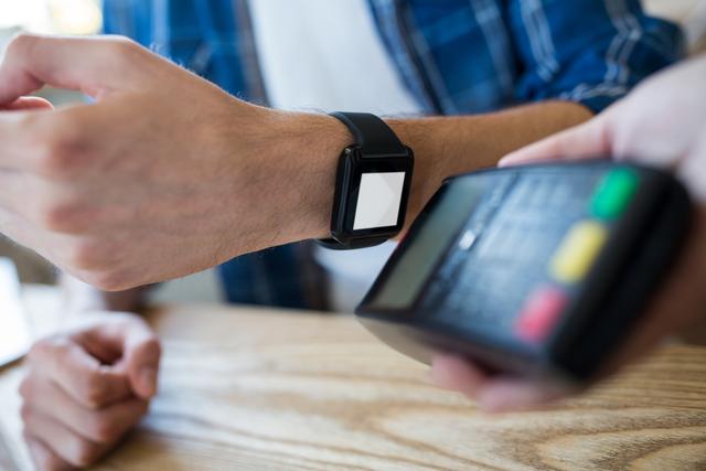 Man making a contactless payment using a smartwatch at a table. Ideal for illustrating modern technology, digital wallets, and the convenience of cashless transactions. Suitable for use in articles, advertisements, and blogs about fintech, wearable technology, and innovative payment solutions.