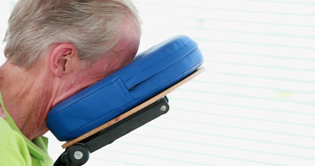 Elderly man lying face down on massage chair with blue cushion. Ideal for articles and materials focusing on elderly care, therapeutic treatments, massage therapy, and relaxation techniques.