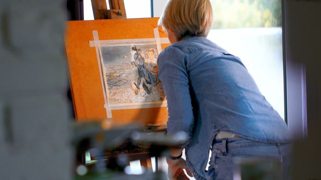 A female artist is engaging in painting on a canvas in her home studio. Her focused expression and dedicated posture highlight her commitment to the art piece she is bringing to life with a paintbrush. This image is ideal for illustrating concepts related to creativity, artistry, home-based creative pursuits, or artistic passion. It can be used in articles, blog posts, or advertising spaces about art education, home studios, or promoting art materials.