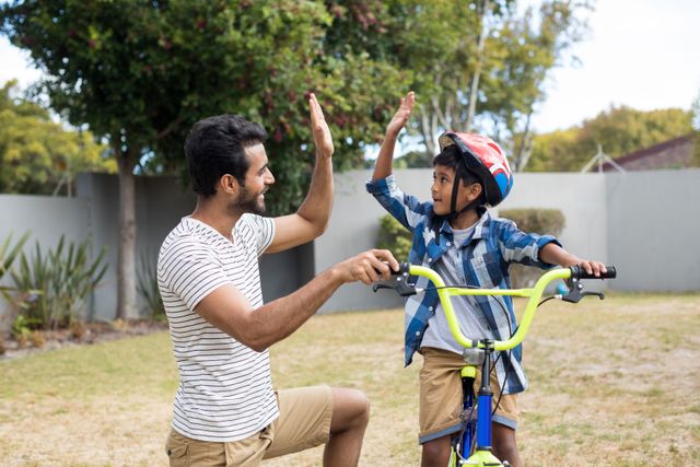 Father and son enjoying a moment of bonding while cycling in the yard. The father is kneeling and giving a high-five to his son, who is wearing a helmet and sitting on a bike. This image can be used for themes related to family bonding, parenting, outdoor activities, and childhood learning experiences.