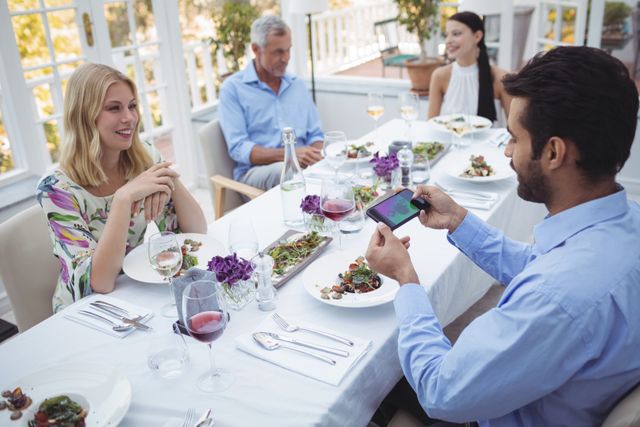 Man taking picture of food with mobile phone while dining with friends and family in a restaurant. Ideal for use in articles about social media trends, dining experiences, food photography, and modern lifestyle.