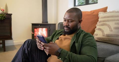 African American Man Relaxing at Home Using Smartphone by Cozy Fireplace