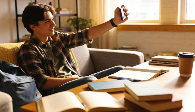 College student taking selfie while studying on couch in sunlit dorm room with coffee