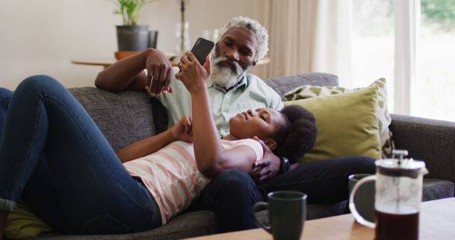 Father and daughter lounging comfortably on the sofa at home, enjoying quality time. Daughter is using a smartphone, with father watching over her shoulder. This image is great for promoting family bonds, home life, and digital connectivity. It can be useful for advertising items related to family activities, parenting, and technology use at home.