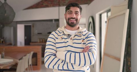 Smiling indian man standing in sunlit modern kitchen studio wearing striped sweater